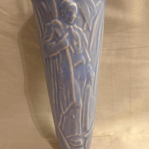 May include: A light blue ceramic wall pocket vase featuring a molded relief design of a figure standing among tall reeds or grasses. The vessel has a tapered, conical shape with a smooth, glossy finish and a wide, open top rim for holding items.
