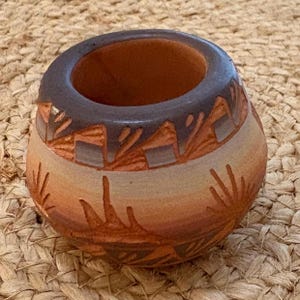 May include: A small, round, hand-painted ceramic pot with a wide opening. The pot features a dark brown rim and a body with a gradient of colors from light tan to reddish-brown. The design includes carved geometric and stylized plant motifs.