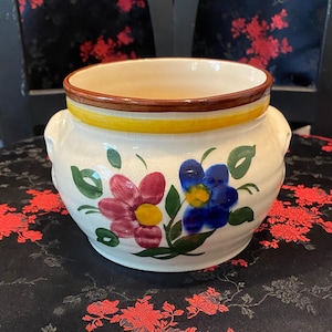May include: A white ceramic bowl with two handles and a floral design. The bowl has a brown rim with a yellow stripe and is decorated with red and blue flowers with green leaves.