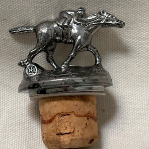 May include: A silver-colored bottle stopper featuring a detailed sculpture of a racehorse with a jockey. The stopper is mounted on a natural cork base, suitable for sealing wine bottles. The design showcases equestrian theme.