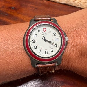 May include: A Swiss Army wristwatch with a white dial, black Arabic numerals, and a red bezel. The watch features a date window at the three o'clock position, black hands, and a brown leather strap. The dial displays the Swiss Army logo.