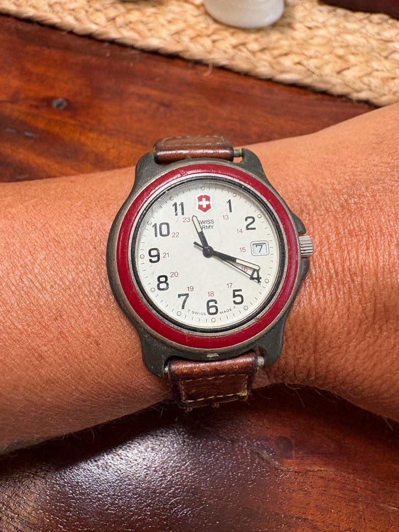 May include: A Swiss Army wristwatch with a white dial, black Arabic numerals, and a red bezel. The watch features a date window at the three o'clock position, black hands, and a brown leather strap. The dial displays the Swiss Army logo.
