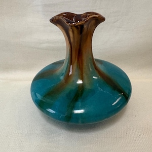 May include: A small, ceramic vase with a round base and a flared, ruffled top. The vase is glazed in a glossy teal blue with brown and bronze accents. The vase has a unique, organic shape.