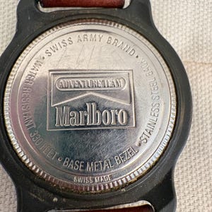 May include: The back of a wristwatch with a brown leather strap and a black case. The stainless steel case back features the text Swiss Army Brand, Adventure Team, Marlboro, Water Resistant 330 Feet, Base Metal Bezel, and Swiss Made.