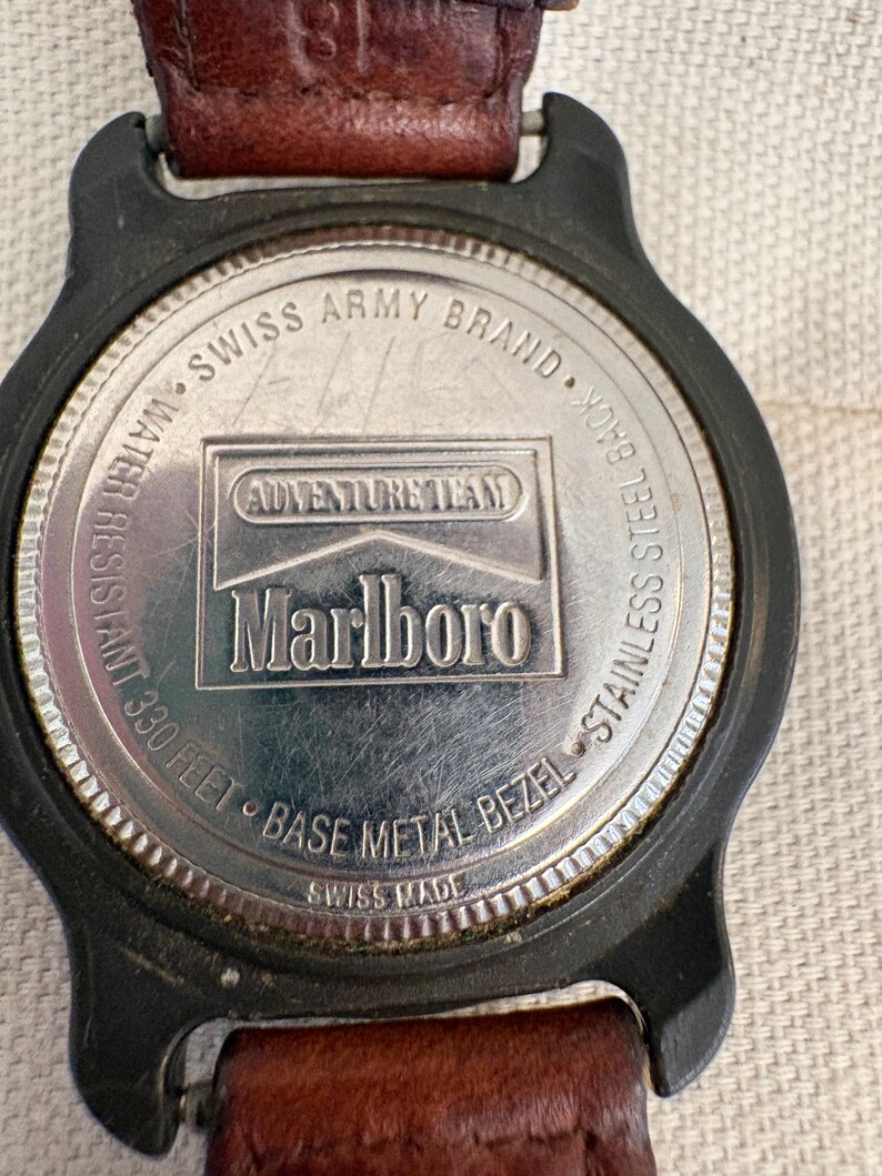 May include: The back of a wristwatch with a brown leather strap and a black case. The stainless steel case back features the text Swiss Army Brand, Adventure Team, Marlboro, Water Resistant 330 Feet, Base Metal Bezel, and Swiss Made.
