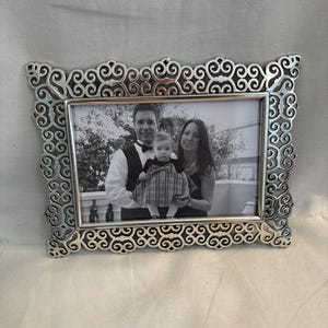 May include: A rectangular silver-toned picture frame featuring an intricate, openwork scrollwork border. The frame holds a black and white photograph of two adults and a small child standing together outdoors in front of a white railing.