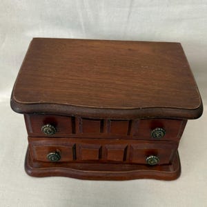 May include: A small wooden chest with a dark brown finish featuring two horizontal drawers. Each drawer is equipped with two circular, metallic knobs with a textured pattern. The chest has a smooth, rectangular top surface and a curved base.