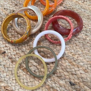 May include: A collection of colorful bangles in various shades of orange, pink, white, and green. Some bangles have decorative elements, such as painted lines or textured surfaces. The bangles are arranged on a woven, tan surface.