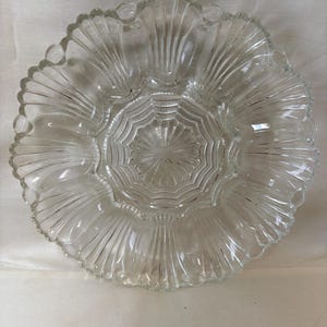 May include: A clear glass serving dish with a scalloped edge and a central starburst design. The dish has multiple compartments and a textured, fan-like pattern radiating from the center. Ideal for serving appetizers or snacks.