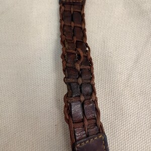 May include: A watch strap made of dark brown leather strips woven with thin brown cord. The strap features a rectangular leather end piece with visible stitching and connects to a dark grey watch casing with a partial red accent at the top.