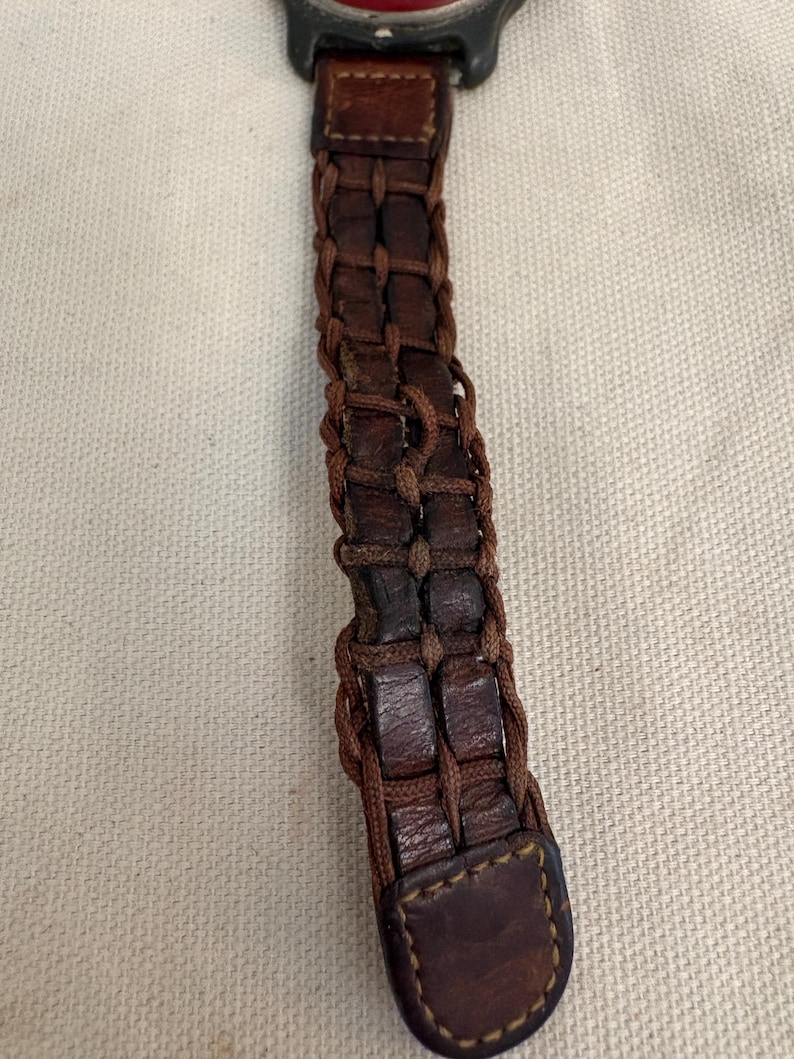 May include: A watch strap made of dark brown leather strips woven with thin brown cord. The strap features a rectangular leather end piece with visible stitching and connects to a dark grey watch casing with a partial red accent at the top.