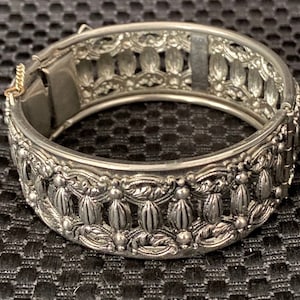 May include: A silver-toned metal hinged bangle bracelet featuring an intricate openwork design with repeating vertical oval elements and decorative scrollwork. The bracelet includes a secure clasp mechanism and a small attached safety chain.