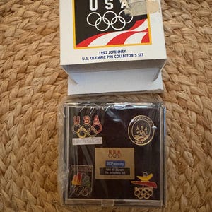 May include: A 1992 JCPenney U.S. Olympic pin collector's set. The box features the USA logo with Olympic rings and a colorful flame. The set includes pins with the USA logo, Olympic rings, and Barcelona '92.