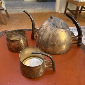 May include: Antique silver tea set with a teapot and two small cups. The teapot has a curved handle and a small knob on the lid. The cups have small handles. The set is on a red surface.