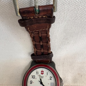 May include: A Swiss Army wristwatch featuring a white dial with black numerals, a red bezel, and a brown leather strap with a woven texture and a silver-toned metal buckle. The watch face displays the Swiss Army logo and a date window at the three o'clock position.