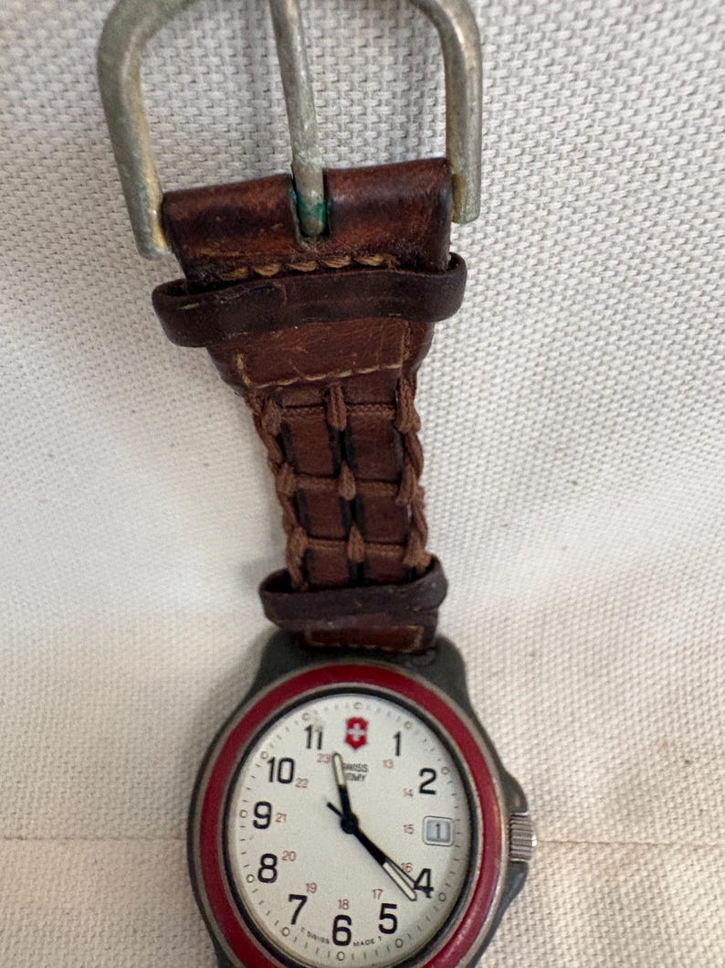 May include: A Swiss Army wristwatch featuring a white dial with black numerals, a red bezel, and a brown leather strap with a woven texture and a silver-toned metal buckle. The watch face displays the Swiss Army logo and a date window at the three o'clock position.