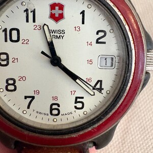May include: A Swiss Army wristwatch with a white dial, black Arabic numerals, and a red bezel. The face features a red shield logo, black hour and minute hands, a date window at the three o'clock position, and a brown leather strap.