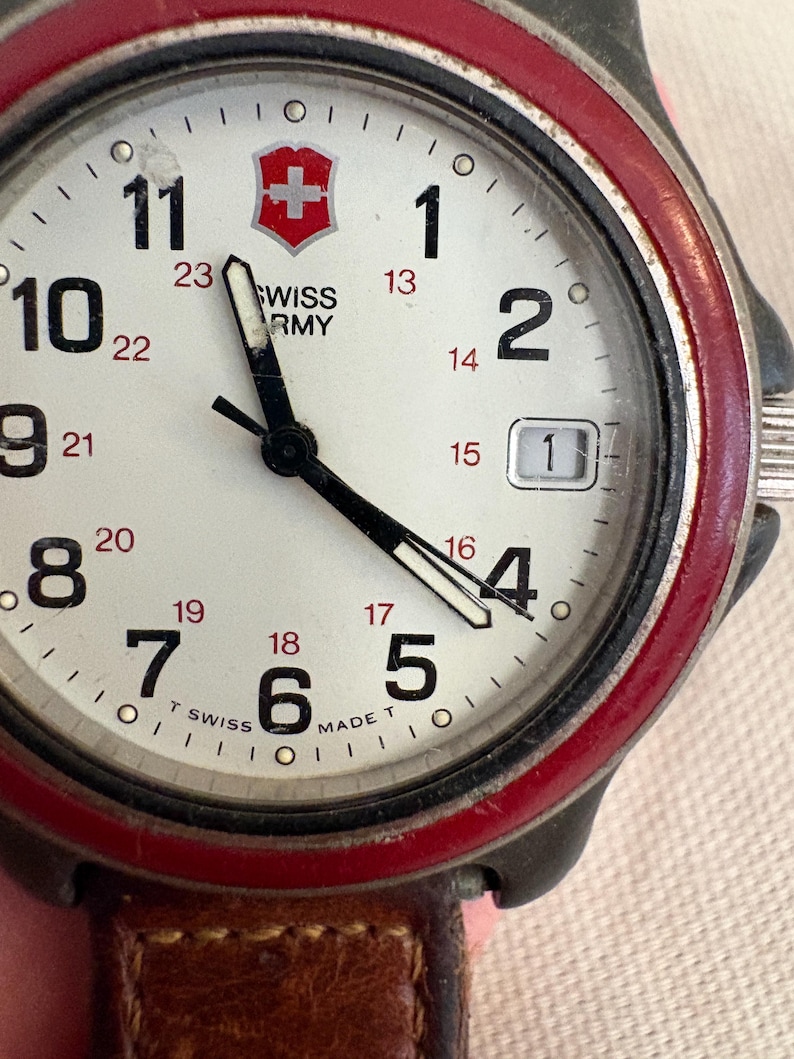 May include: A Swiss Army wristwatch with a white dial, black Arabic numerals, and a red bezel. The face features a red shield logo, black hour and minute hands, a date window at the three o'clock position, and a brown leather strap.