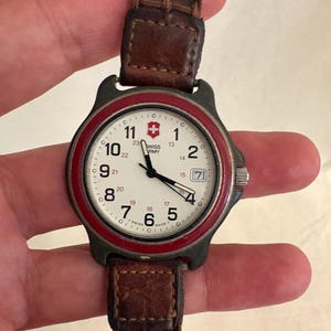 May include: A Swiss Army wristwatch with a cream-colored dial, black Arabic numerals, a red bezel, and a dark brown braided leather strap. The watch face features a date window at the three o'clock position and the Swiss Army logo at the top.