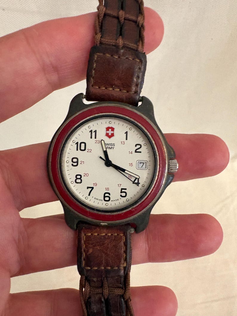 May include: A Swiss Army wristwatch with a cream-colored dial, black Arabic numerals, a red bezel, and a dark brown braided leather strap. The watch face features a date window at the three o'clock position and the Swiss Army logo at the top.
