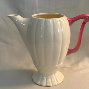 May include: A cream-colored ceramic pitcher featuring a vertical ribbed pattern on the body, a flared pedestal base, and a bright pink handle. The pitcher has a pointed spout and a smooth, glossy finish, standing upright with a wide opening.