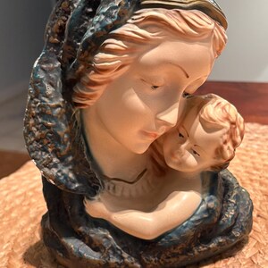 May include: A ceramic figurine depicting a mother and child in a close embrace. The mother wears a textured, dark teal head covering with gold accents, while the child is nestled against her. The figures have light skin tones and soft features.