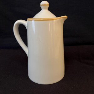 May include: A white ceramic pitcher with a lid. The pitcher has a curved handle and a spout. The lid and the rim of the pitcher are trimmed with a thin band of tan. The pitcher is on a dark background.