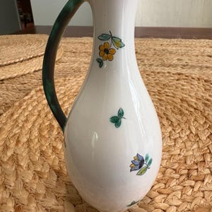 May include: White ceramic pitcher with a green handle and rim, adorned with hand-painted yellow and blue floral designs. The pitcher has a slender neck and a curved handle, perfect for serving beverages.