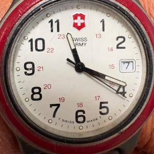 May include: A Swiss Army wristwatch with a white dial, black numerals, and a red bezel. The face features a red shield logo, the text SWISS ARMY, and a date window showing the number 7. It has black hour, minute, and second hands with luminous inserts.