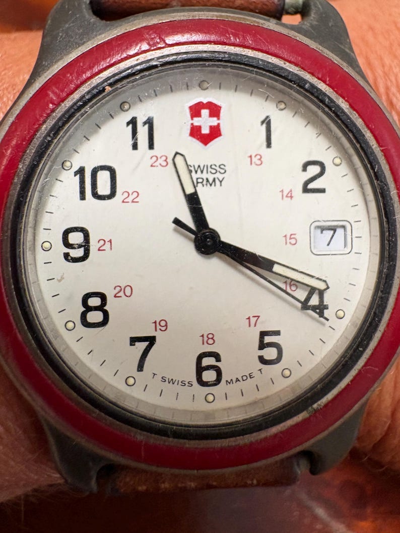 May include: A Swiss Army wristwatch with a white dial, black numerals, and a red bezel. The face features a red shield logo, the text SWISS ARMY, and a date window showing the number 7. It has black hour, minute, and second hands with luminous inserts.