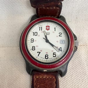 May include: A Swiss Army wristwatch with a white dial, black Arabic numerals, and a red bezel. The watch features a date window at the three o'clock position, black hands, and a brown leather strap with visible stitching and a metal buckle.