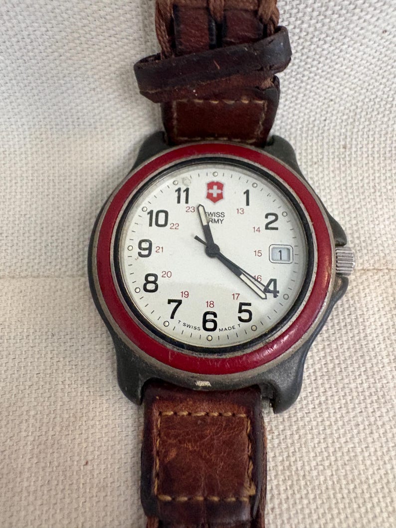 May include: A Swiss Army wristwatch with a white dial, black Arabic numerals, and a red bezel. The watch features a date window at the three o'clock position, black hands, and a brown leather strap with visible stitching and a metal buckle.