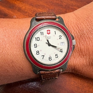 May include: A Swiss Army wristwatch with a cream-colored dial, black Arabic numerals, and a red bezel. The watch features a date window at the three o'clock position, black hands, and a brown leather strap. The dial displays the Swiss Army logo.