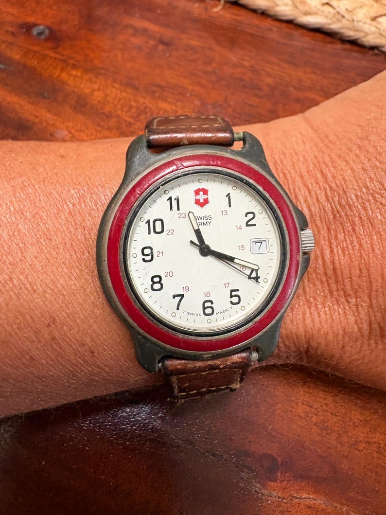 May include: A Swiss Army wristwatch with a cream-colored dial, black Arabic numerals, and a red bezel. The watch features a date window at the three o'clock position, black hands, and a brown leather strap. The dial displays the Swiss Army logo.