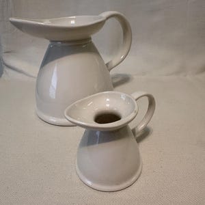 May include: Two white ceramic pitchers. The larger pitcher has a wide, flared spout and a curved handle. The smaller pitcher mirrors the design, with a similar spout and handle. Both are on a neutral background.