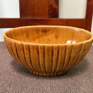 May include: A round ceramic bowl with a glossy, mottled amber-brown glaze. The exterior features a series of vertical, evenly spaced fluted ridges that extend from the base to the rim, creating a textured, scalloped appearance around the circumference.