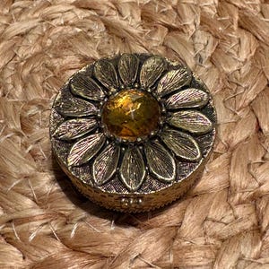 May include: A round, antique-style brass trinket box featuring a lid decorated with a dimensional flower motif. The center of the flower is set with a faceted, amber-colored glass stone, while the petals exhibit a textured, metallic finish.