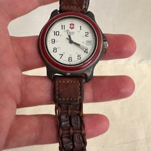 May include: A Swiss Army wristwatch with a white dial, black Arabic numerals, and a red bezel. The watch features a date window at the three o'clock position and a brown braided leather strap. The dial displays the Swiss Army logo and text.