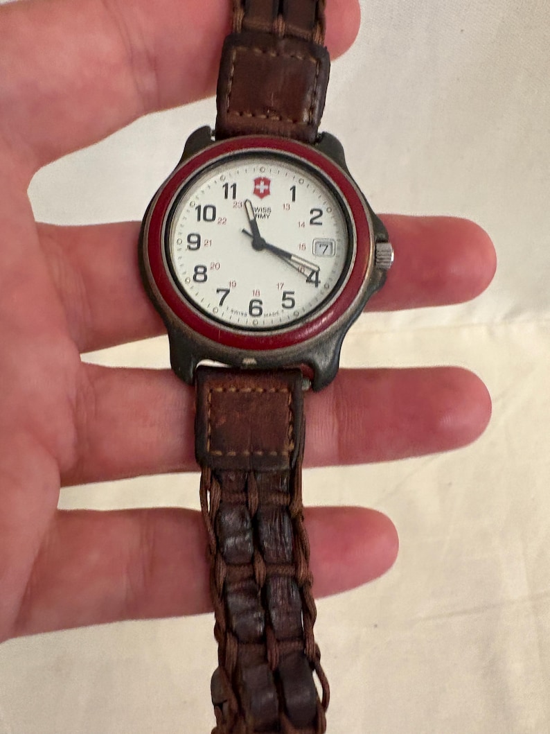 May include: A Swiss Army wristwatch with a white dial, black Arabic numerals, and a red bezel. The watch features a date window at the three o'clock position and a brown braided leather strap. The dial displays the Swiss Army logo and text.