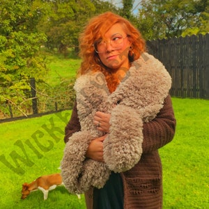 May include: A brown crochet cardigan with a fluffy, light brown collar. The cardigan has pockets and is worn over a dark skirt. The background features a green lawn, trees, and a dog.