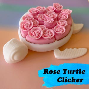 May include: A white and pink rose turtle clicker. The turtle's shell is covered in pink roses. The clicker has white flippers and a white head. The text "Rose Turtle Clicker" is in a blue rectangle.