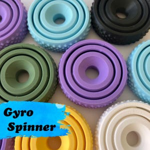 May include: A collection of circular, multi-layered gyro spinners in various colours including light blue, black, olive green, purple, yellow, and white. Each spinner features a textured, knurled outer edge and a central hole, with the text Gyro Spinner.