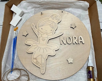 May include: A wooden craft kit featuring a ballerina design with the name "Nora" painted on it. The kit includes paint, brushes, and a sealant.