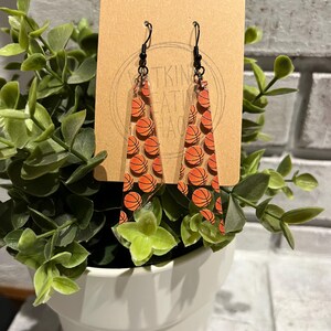 May include: A pair of dangle earrings with a clear acrylic triangle shape. The earrings are decorated with a basketball pattern. The earrings are hanging from a brown card with the text "Witkin Creations".