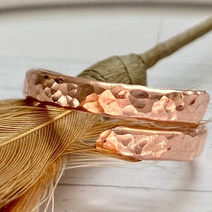 May include: Two hammered copper rings, one slightly larger than the other, are stacked on top of each other. The rings are a warm rose gold color and have a textured, hammered finish.