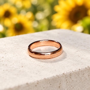 May include: A polished rose gold ring with a smooth, rounded band and a high-shine metallic finish. The ring features a simple, continuous circular design with a reflective surface that catches the light, showcasing its warm, pinkish-gold hue.