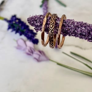 May include: Three thin copper rings stacked on a dried lavender stem. The top ring has a decorative swirl pattern.
