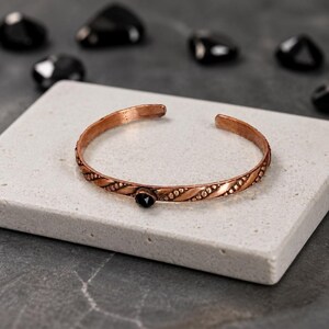 May include: A copper cuff bracelet with a black gemstone. The bracelet has a textured, braided design and is displayed on a white stone surface. Black stones are scattered in the background. Jewelry.