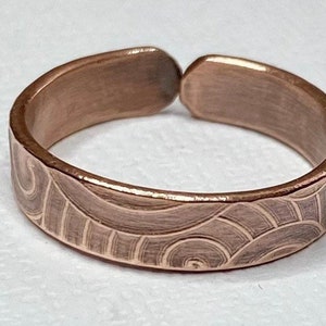 May include: A copper-colored adjustable ring with an engraved wave pattern. The open design allows for size adjustment. The ring's surface has a brushed texture, enhancing the visual appeal of the jewelry.