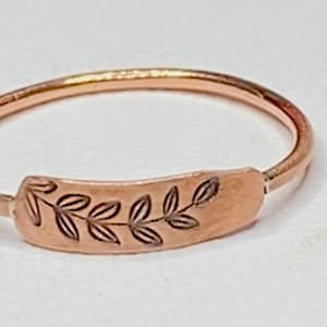 May include: A delicate rose gold ring with a thin band and a rectangular design featuring a leafy branch engraved on the top.